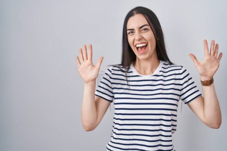 Foto de Joven morena vistiendo camiseta a rayas mostrando y apuntando hacia arriba con los dedos número diez mientras sonríe confiado y feliz. - Imagen libre de derechos