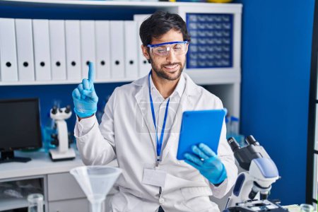 Foto de Hombre latino guapo trabajando en el laboratorio científico usando tableta sonriendo con una idea o pregunta señalando el dedo con la cara feliz, número uno - Imagen libre de derechos