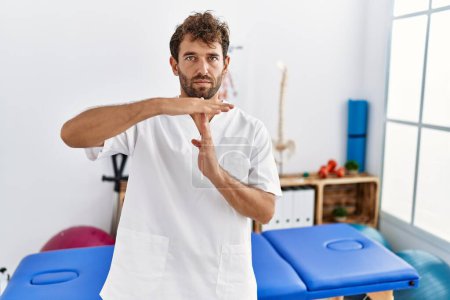 Photo for Young handsome physiotherapist man working at pain recovery clinic doing time out gesture with hands, frustrated and serious face - Royalty Free Image