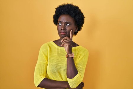 Foto de African young woman standing over yellow studio thinking worried about a question, concerned and nervous with hand on chin - Imagen libre de derechos