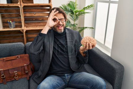 Foto de Guapo hombre de mediana edad en la oficina de consulta sosteniendo el cerebro sonriendo feliz haciendo signo bien con la mano en el ojo mirando a través de los dedos - Imagen libre de derechos