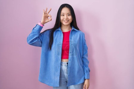 Foto de Joven mujer asiática de pie sobre fondo rosa sonriendo positiva haciendo signo ok con la mano y los dedos. expresión exitosa. - Imagen libre de derechos