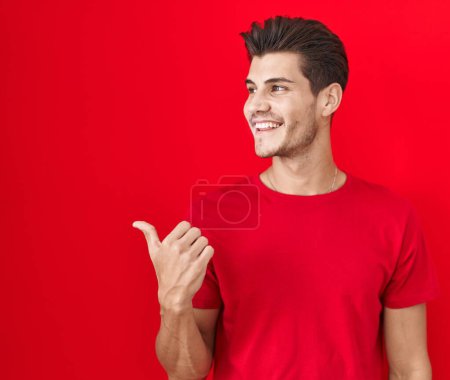 Photo for Young hispanic man standing over red background smiling with happy face looking and pointing to the side with thumb up. - Royalty Free Image