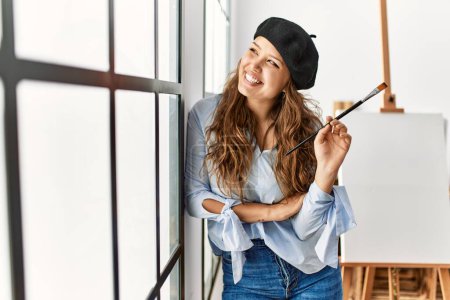Foto de Joven mujer hispana hermosa artista sosteniendo pincel apoyado en la pared en el estudio de arte - Imagen libre de derechos