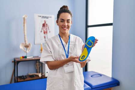 Photo for Young hispanic woman holding shoe insole at physiotherapy clinic winking looking at the camera with sexy expression, cheerful and happy face. - Royalty Free Image