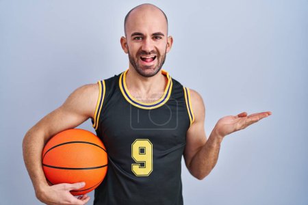 Téléchargez les photos : Jeune homme chauve avec barbe portant l'uniforme de basket tenant ballon pointant de côté avec les mains ouvertes paumes montrant l'espace de copie, présentant la publicité souriant heureux excité - en image libre de droit