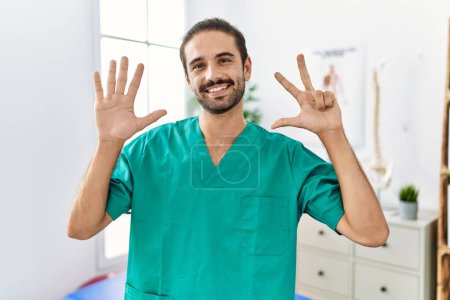 Foto de Joven fisioterapeuta trabajando en la clínica de recuperación del dolor mostrando y apuntando hacia arriba con los dedos número ocho mientras sonríe confiado y feliz. - Imagen libre de derechos
