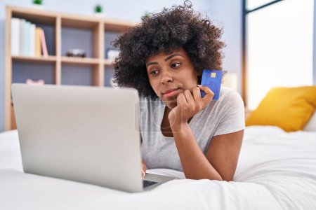Foto de Mujer afroamericana usando laptop y tarjeta de crédito con expresión de duda en el dormitorio - Imagen libre de derechos