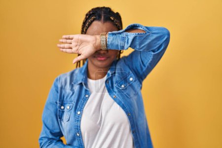 Téléchargez les photos : Femme afro-américaine avec des tresses debout sur fond jaune couvrant les yeux avec le bras, l'air grave et triste. concept de non-visibilité, de dissimulation et de rejet - en image libre de droit