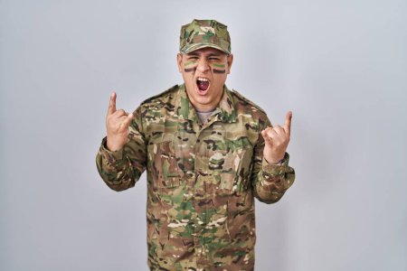 Téléchargez les photos : Jeune homme hispanique portant uniforme de camouflage de l'armée criant avec une expression folle faire symbole rock avec les mains en l'air. star de la musique. concept lourd. - en image libre de droit