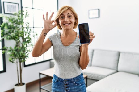 Foto de Mujer rubia de mediana edad que trabaja sosteniendo el teléfono inteligente que muestra la pantalla haciendo signo bien con los dedos, sonriendo gesto amistoso excelente símbolo - Imagen libre de derechos