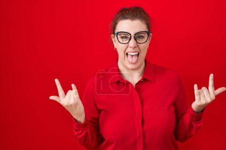 Foto de Young hispanic woman with red hair standing over red background shouting with crazy expression doing rock symbol with hands up. music star. heavy concept. - Imagen libre de derechos