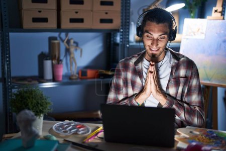 Téléchargez les photos : Jeune homme hispanique assis au studio d'art avec ordinateur portable tard dans la nuit priant avec les mains ensemble demandant pardon souriant confiant. - en image libre de droit