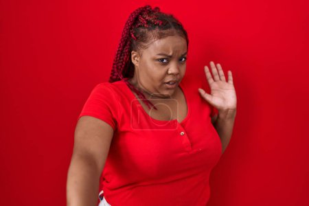 Foto de Mujer afroamericana con el pelo trenzado de pie sobre el fondo rojo asustado y aterrorizado con la expresión del miedo detener el gesto con las manos, gritando en shock. concepto de pánico. - Imagen libre de derechos