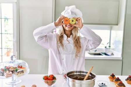 Foto de Young woman wearing cook uniform holding doughnuts over eyes at kitchen - Imagen libre de derechos