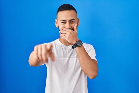 Téléchargez les photos : Jeune homme hispanique debout sur fond bleu se moquant de vous, pointant du doigt la caméra avec la main sur la bouche, expression de honte - en image libre de droit