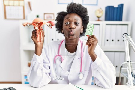 Foto de Mujer doctora africana sosteniendo órganos genitales femeninos anatómicos y píldoras anticonceptivas mirando a la cámara soplando un beso siendo encantadora y sexy. expresión de amor. - Imagen libre de derechos