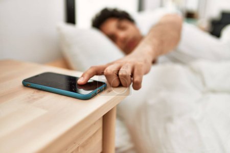 Foto de Hombre hispano joven apagando la alarma del teléfono inteligente acostado en la cama en el dormitorio. - Imagen libre de derechos