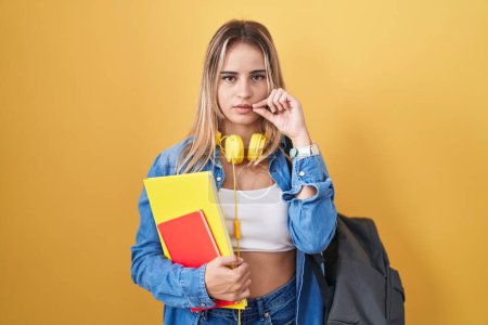 Téléchargez les photos : Young blonde woman wearing student backpack and holding books mouth and lips shut as zip with fingers. secret and silent, taboo talking - en image libre de droit