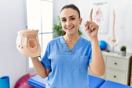 Photo for Young physiotherapist woman holding cervical neck collar at medical clinic surprised with an idea or question pointing finger with happy face, number one - Royalty Free Image
