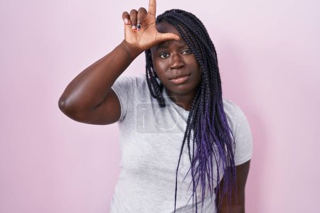 Foto de Joven mujer africana de pie sobre fondo rosa burlándose de la gente con los dedos en la frente haciendo gesto perdedor burlándose e insultando. - Imagen libre de derechos