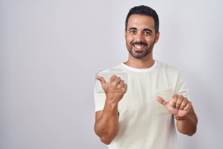 Téléchargez les photos : Homme hispanique avec la barbe debout sur fond isolé pointant vers le dos derrière avec la main et les pouces vers le haut, souriant confiant - en image libre de droit