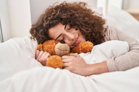 Photo for Young hispanic woman hugging teddy bear lying on bed sleeping at bedroom - Royalty Free Image