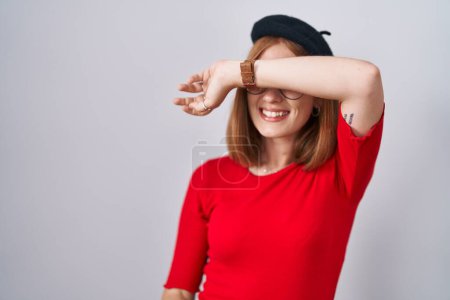 Foto de Mujer pelirroja joven de pie con gafas y boina cubriendo los ojos con el brazo sonriendo alegre y divertido. concepto ciego. - Imagen libre de derechos