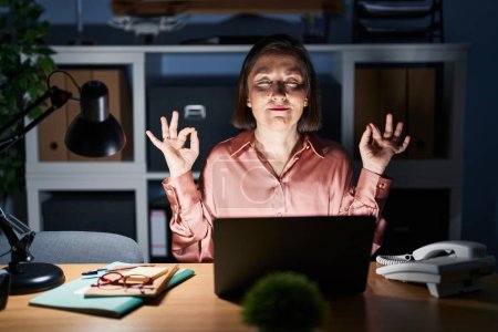 Téléchargez les photos : Femme hispanique du Moyen Âge travaillant à l'aide d'un ordinateur portable tard dans la nuit se détendre et sourire les yeux fermés en faisant geste de méditation avec les doigts. concept de yoga. - en image libre de droit
