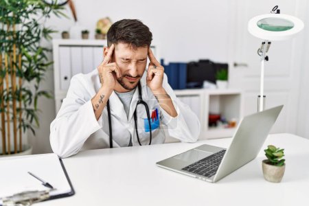 Foto de Joven médico que trabaja en la clínica usando computadora portátil con la mano en la cabeza para el dolor en la cabeza porque el estrés. padecer migraña. - Imagen libre de derechos