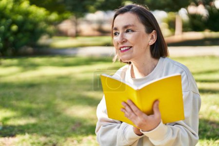 Téléchargez les photos : Femme d'âge moyen lecture livre assis sur herbe au parc - en image libre de droit