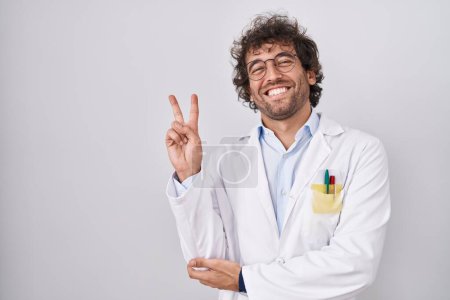 Foto de Joven hispano vistiendo uniforme médico sonriendo con la cara feliz guiñando un ojo a la cámara haciendo señal de victoria. número dos. - Imagen libre de derechos