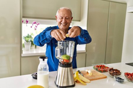 Téléchargez les photos : Senior homme souriant confiant mettre la banane dans le mélangeur à la cuisine - en image libre de droit