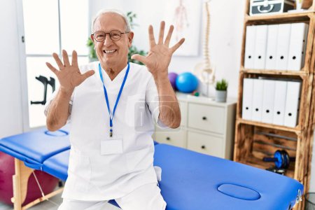 Téléchargez les photos : Homme de physiothérapie senior travaillant à la clinique de récupération de la douleur montrant et pointant vers le haut avec les doigts numéro dix tout en souriant confiant et heureux. - en image libre de droit