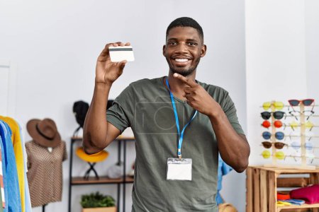 Photo for Young african american man working as manager at retail boutique holding credit card smiling happy pointing with hand and finger - Royalty Free Image