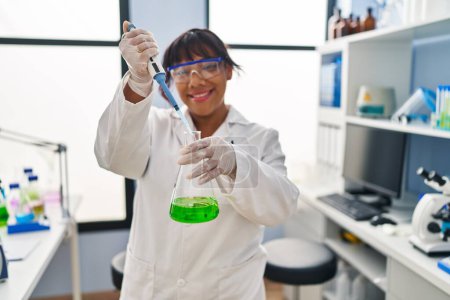 Téléchargez les photos : Jeune belle femme latine scientifique souriant confiant versant du liquide sur éprouvette au laboratoire - en image libre de droit