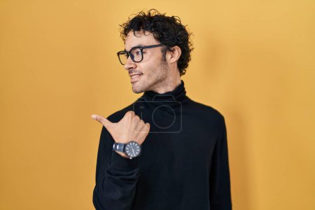 Photo for Hispanic man standing over yellow background smiling with happy face looking and pointing to the side with thumb up. - Royalty Free Image