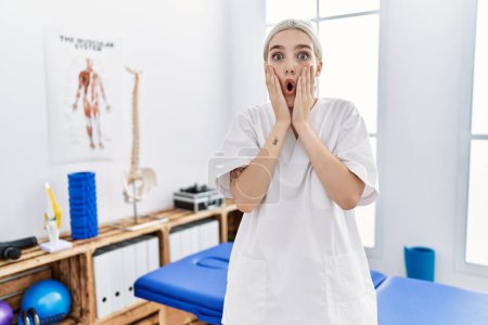 Foto de Mujer caucásica joven que trabaja en la clínica de recuperación del dolor asustado y sorprendido, sorpresa y expresión asombrada con las manos en la cara - Imagen libre de derechos