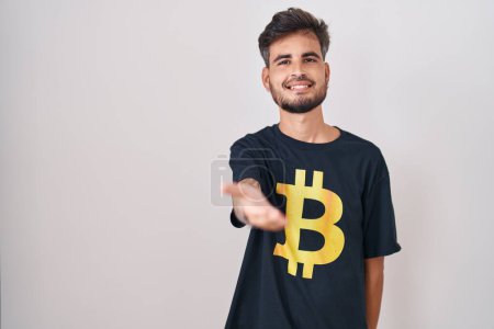 Photo for Young hispanic man with tattoos wearing bitcoin t shirt smiling cheerful offering palm hand giving assistance and acceptance. - Royalty Free Image