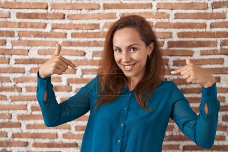 Foto de Brunette woman standing over bricks wall looking confident with smile on face, pointing oneself with fingers proud and happy. - Imagen libre de derechos