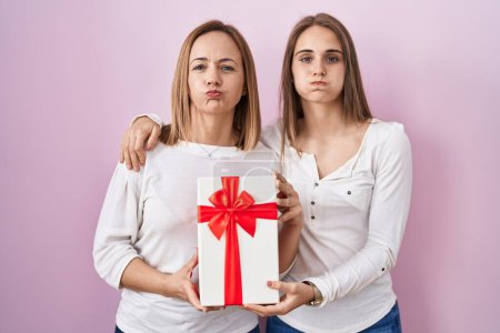 Foto de Middle age mother and young daughter holding mothers day gift puffing cheeks with funny face. mouth inflated with air, catching air. - Imagen libre de derechos