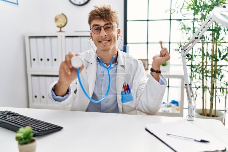 Foto de Young caucasian doctor man wearing doctor uniform and using stethoscope at the clinic screaming proud, celebrating victory and success very excited with raised arm - Imagen libre de derechos