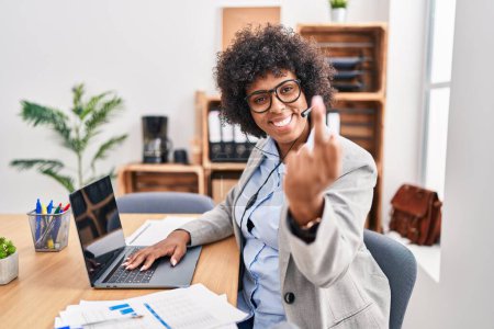 Foto de Black woman with curly hair wearing call center agent headset at the office showing middle finger, impolite and rude fuck off expression - Imagen libre de derechos