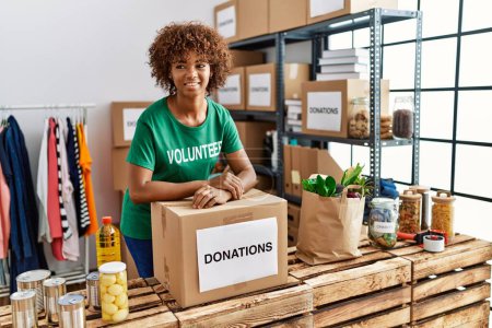 Téléchargez les photos : Young african american woman wearing volunteer uniform leaning on donations box at charity center - en image libre de droit