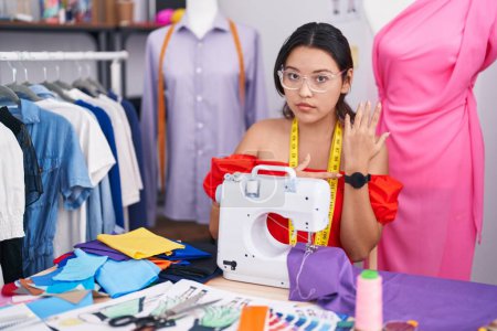 Photo for Hispanic young woman dressmaker designer using sewing machine in hurry pointing to watch time, impatience, looking at the camera with relaxed expression - Royalty Free Image