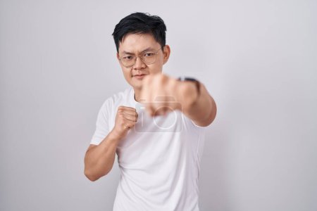 Foto de Joven asiático de pie sobre fondo blanco puñetazo puño para luchar, ataque agresivo y enojado, amenaza y violencia - Imagen libre de derechos
