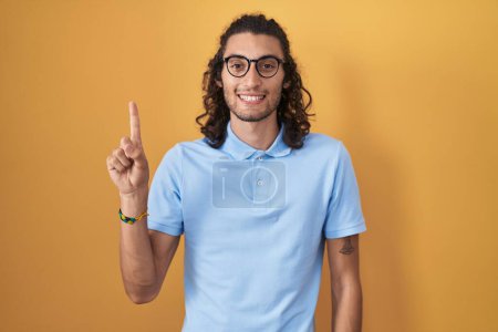 Photo for Young hispanic man standing over yellow background showing and pointing up with finger number one while smiling confident and happy. - Royalty Free Image