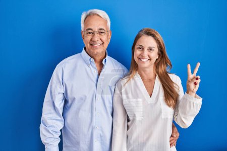 Photo for Middle age hispanic couple standing over blue background showing and pointing up with fingers number two while smiling confident and happy. - Royalty Free Image