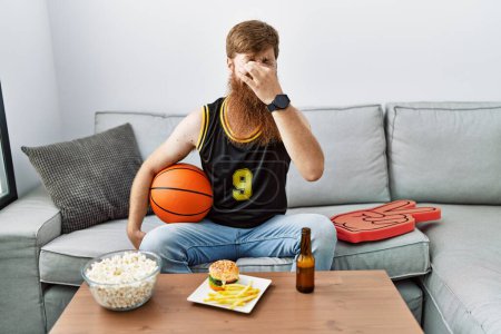Photo for Caucasian man with long beard holding basketball ball cheering tv game smelling something stinky and disgusting, intolerable smell, holding breath with fingers on nose. bad smell - Royalty Free Image