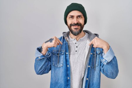 Foto de Joven hombre hispano con tatuajes con gorra de lana mirando confiado con sonrisa en la cara, señalándose con los dedos orgullosos y felices. - Imagen libre de derechos